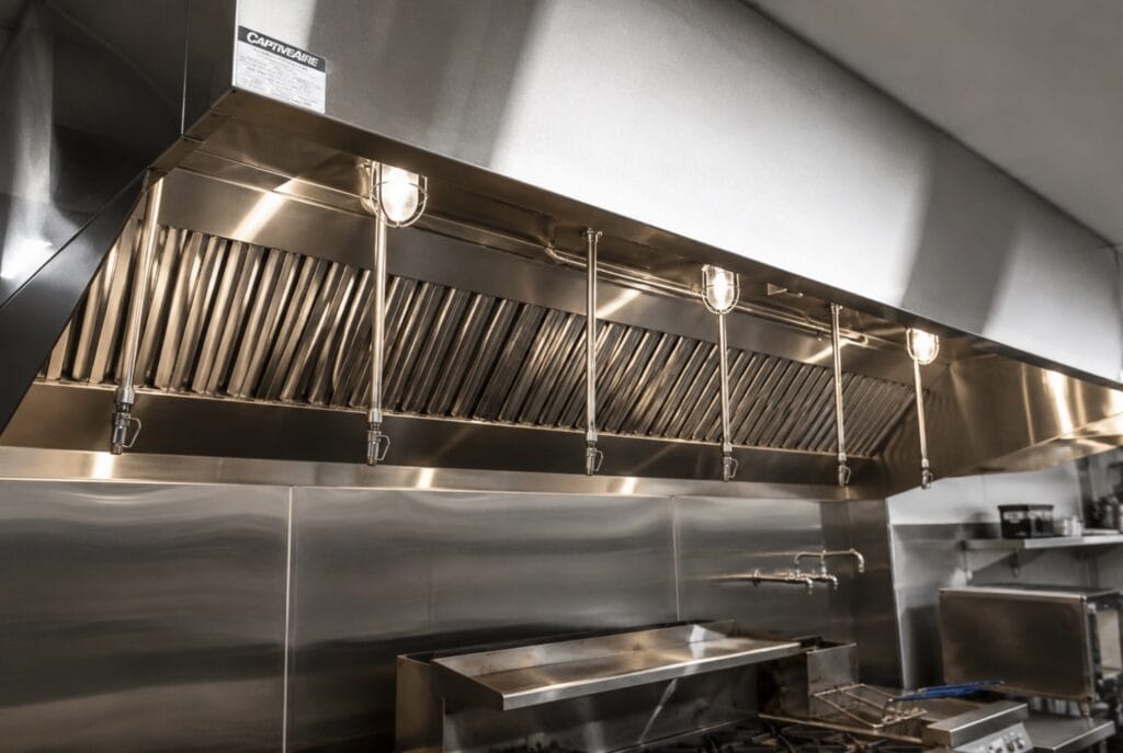 Commercial Type I hood and exhaust system installed over a restaurant cookline with stainless steel wall panels and kitchen equipment.