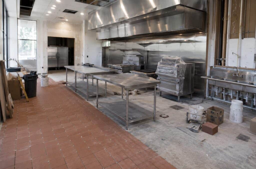Restaurant kitchen renovation in progress with stainless prep tables, hood system, unfinished walls, and floor tile partially installed.